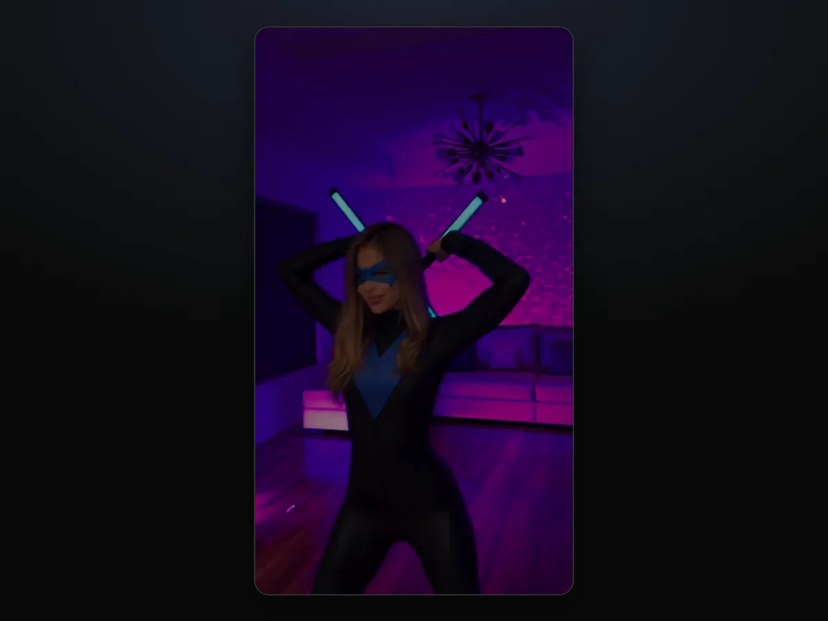 Fotograma de un video del trend de Nightwing IA hecho en TryViralAI: una mujer joven con el traje negro con la V azul y la máscara azul, con los dos bastones luminosos cruzados detrás de la cabeza